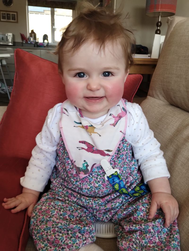 A baby sat up on the sofa with a red cushion tucked behind them.