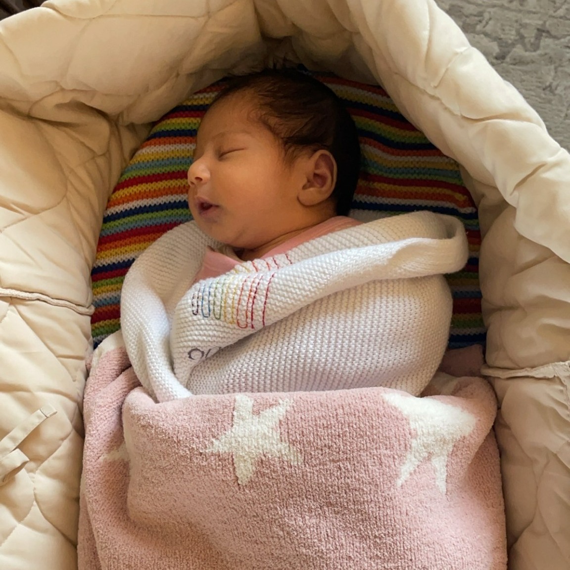 A baby sleeping in a moses basket wrapped in a white and pink blanket.