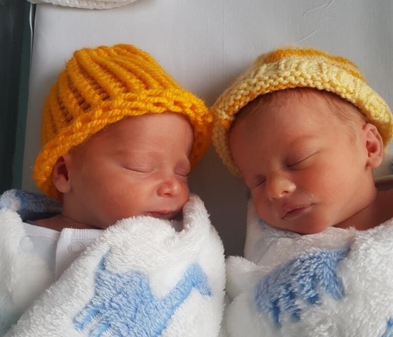 Two babies in a bed together wearing knitted hats and both wrapped in a white and blue blanket.