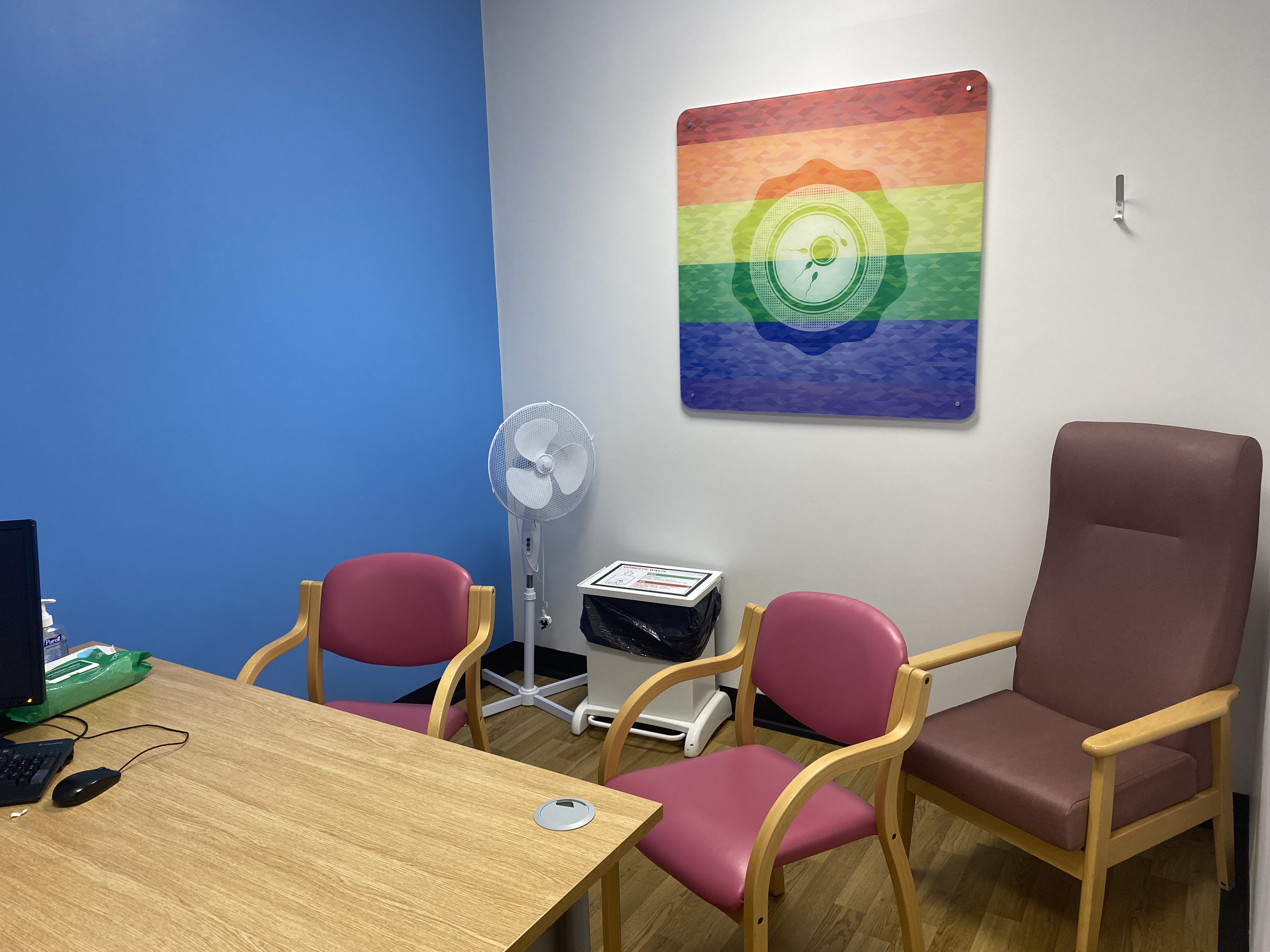 A room with a blue wall and a white wall, with a picture frame. A desk, 3 chairs, and a fan