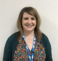A person with long blonde hair wearing a black cardigan and flowery top and blue lanyard.