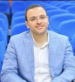 Man wearing glasses and a light blue blazer, seated in front of rows of blue auditorium seats, smiling at the camera.