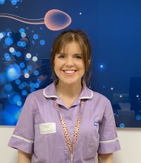 A person smiling with long brunette hair, and a purple uniform and rainbow lanyard.
