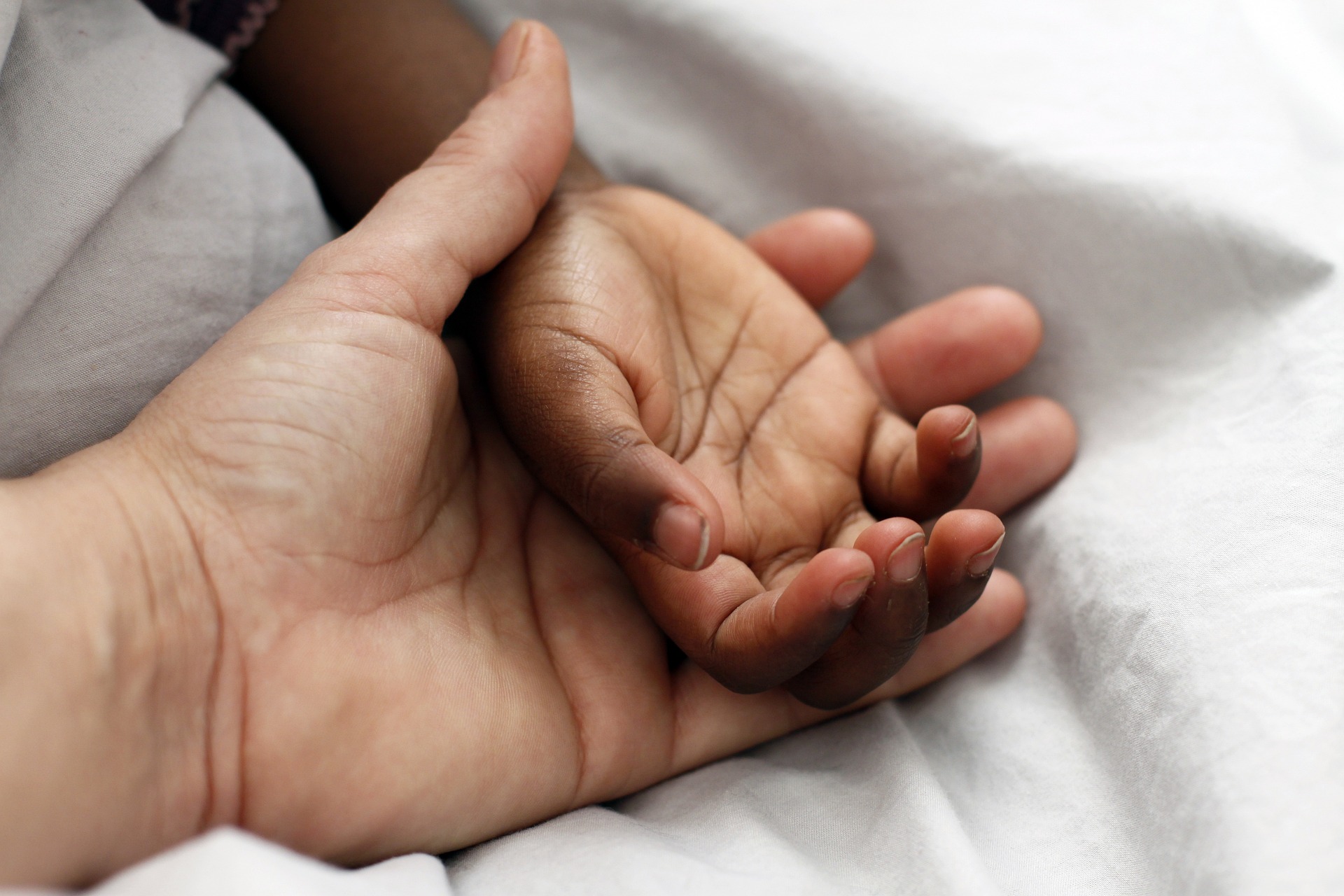A close-up of an adult's hand holding a small hand on top of a white cover.